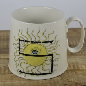 Pode incluir: Caneca de cer&acirc;mica branca com um contorno preto da letra "S" e um sol amarelo com um olho azul no centro. O sol tem raios amarelos.