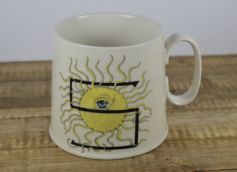 Pode incluir: Caneca de cer&acirc;mica branca com um contorno preto da letra "S" e um sol amarelo com um olho azul no centro. O sol tem raios amarelos.