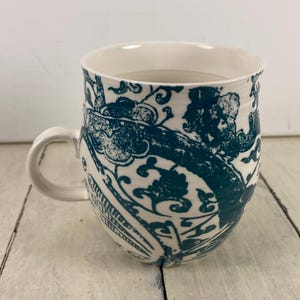 K&ouml;nnte beinhalten: Wei&szlig;er Keramikbecher mit einem tealfarbenen Blumenmuster. Der Becher hat eine abgerundete Form, einen gebogenen Henkel und eine breite &Ouml;ffnung. Das tealfarbene Design bedeckt die Au&szlig;enseite und schafft ein dekoratives und praktisches Trinkgef&auml;&szlig;.