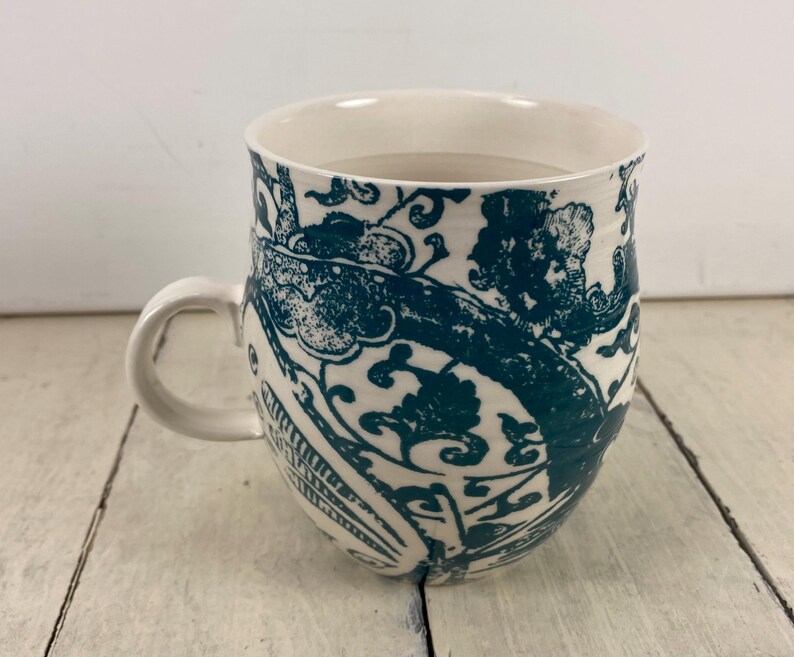 K&ouml;nnte beinhalten: Wei&szlig;er Keramikbecher mit einem tealfarbenen Blumenmuster. Der Becher hat eine abgerundete Form, einen gebogenen Henkel und eine breite &Ouml;ffnung. Das tealfarbene Design bedeckt die Au&szlig;enseite und schafft ein dekoratives und praktisches Trinkgef&auml;&szlig;.