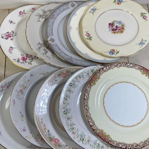 May include: A collection of nine ceramic dinner plates arranged in two rows. Each plate features distinct floral patterns, gold or silver rim detailing, and scalloped or smooth edges. The designs include various pastel colors, intricate botanical motifs, and decorative borders on white or cream-colored backgrounds.