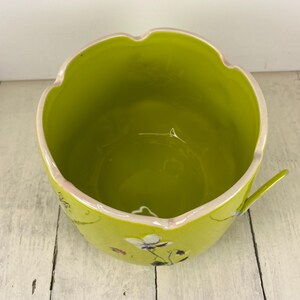 Może przedstawiać: Jasnozielona ceramiczna miska z falistym brzegiem i małym uchwytem. Miska ma białe wnętrze i jest ozdobiona delikatnymi wzorami kwiatowymi w kolorze białym i fioletowym. Miska stoi na białej drewnianej powierzchni.