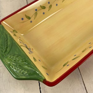 May include: A rectangular ceramic serving tray with a cream-colored center, a deep red rim, and green handles featuring embossed wheat stalks. The interior surface is decorated with a painted vine pattern of green leaves and small blue dots.