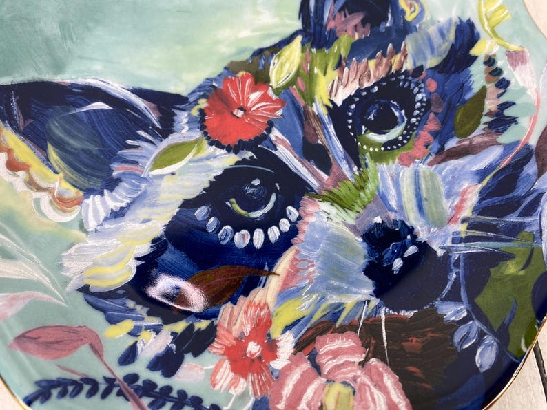 May include: A decorative ceramic plate featuring a colorful floral design with a raccoon face. The plate has a light blue background with red, pink, blue, and yellow flowers. The rim is gold.