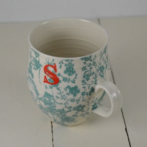 May include: White ceramic mug with a teal floral pattern and a red letter "S" on the side. The mug has a rounded shape and a handle.