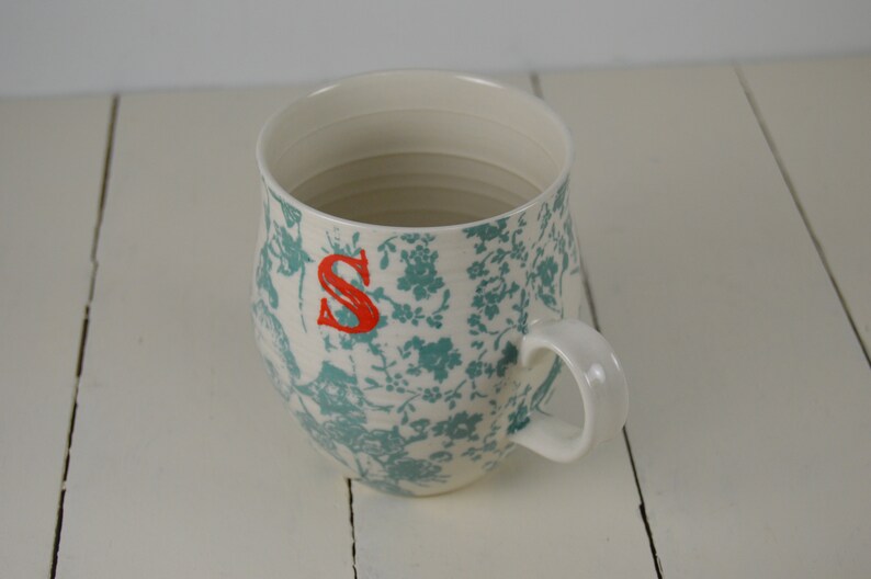 May include: White ceramic mug with a teal floral pattern and a red letter "S" on the side. The mug has a rounded shape and a handle.