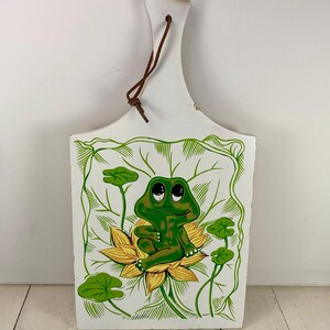May include: White wooden cutting board with a hand-painted frog design. The frog is green and sits on a yellow flower, surrounded by green leaves. A brown leather strap is attached at the top for hanging. The board is approximately 12 inches long.