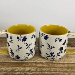 May include: Two ceramic mugs with a cream base and a navy floral pattern. The interior and handle are yellow. The handle has a geometric design. The mugs are on a wooden surface.