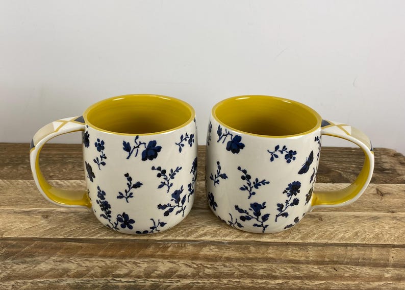 May include: Two ceramic mugs with a cream base and a navy floral pattern. The interior and handle are yellow. The handle has a geometric design. The mugs are on a wooden surface.