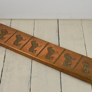 May include: A long, rectangular copper baking pan with a raised design of seven lions. The pan is approximately 18 inches long and 3 inches wide.