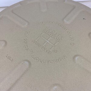 May include: Tan stoneware baking dish with embossed text: "THE PAMPERED CHEF FAMILY HERITAGE STONEWARE CLASSICS COLLECTION USA." The dish has a textured surface and a circular shape. The number "7" is also embossed.