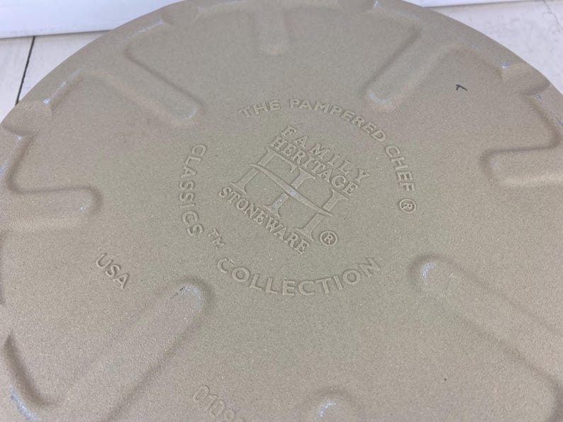 May include: Tan stoneware baking dish with embossed text: "THE PAMPERED CHEF FAMILY HERITAGE STONEWARE CLASSICS COLLECTION USA." The dish has a textured surface and a circular shape. The number "7" is also embossed.