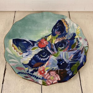 May include: A decorative ceramic plate featuring a colorful raccoon design. The plate has a scalloped edge and a gold rim. The raccoon is painted with blue, white, and pink hues, surrounded by floral accents. The background is a soft teal.