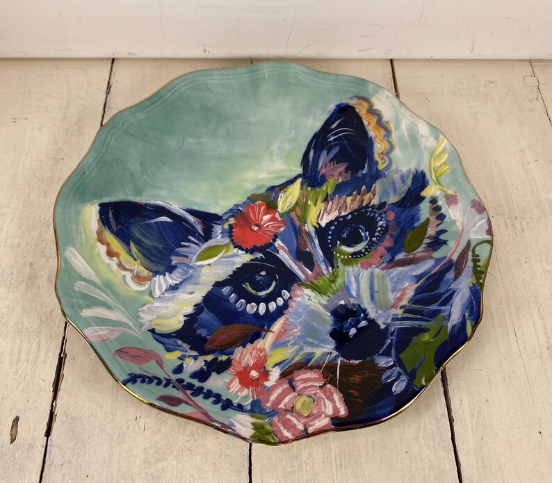 May include: A decorative ceramic plate featuring a colorful raccoon design. The plate has a scalloped edge and a gold rim. The raccoon is painted with blue, white, and pink hues, surrounded by floral accents. The background is a soft teal.