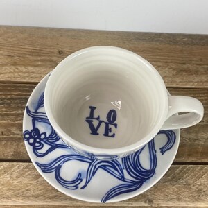 Puede incluir: Juego de taza y platillo de t&eacute; de cer&aacute;mica blanca. La taza tiene un asa y la palabra "LOVE" en azul en el interior. El platillo presenta un dise&ntilde;o floral azul. El juego est&aacute; sobre una superficie de madera.