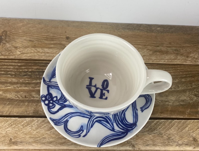 Puede incluir: Juego de taza y platillo de t&eacute; de cer&aacute;mica blanca. La taza tiene un asa y la palabra "LOVE" en azul en el interior. El platillo presenta un dise&ntilde;o floral azul. El juego est&aacute; sobre una superficie de madera.