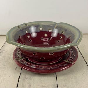 May include: A handmade ceramic colander and matching plate set. The colander is a deep burgundy color with a light green rim and numerous drainage holes. The plate is burgundy with a decorative wavy edge. A unique kitchenware set.