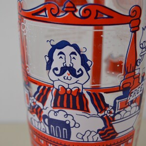 May include: A vintage glass mixing pitcher with a red, white, and blue cartoon illustration of a man with a mustache holding a beer mug. The pitcher has a red rim and a red handle. The text "FREE" is visible on the pitcher.