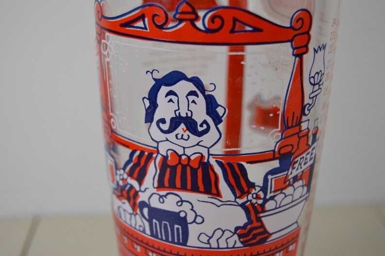 May include: A vintage glass mixing pitcher with a red, white, and blue cartoon illustration of a man with a mustache holding a beer mug. The pitcher has a red rim and a red handle. The text "FREE" is visible on the pitcher.