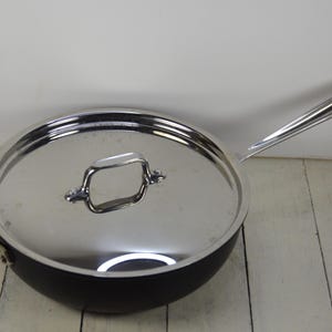 May include: A stainless steel saucepan with a black handle and a lid. The lid has a handle on top.