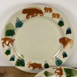 May include: Cream-colored ceramic plate with a forest animal design. The plate features brown bears, a deer, a moose, and a bison, along with green and blue trees. The plate is round and has a wide rim.