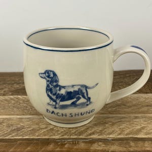 K&ouml;nnte beinhalten: Cremefarbene Keramiktasse mit einer blauen Dackel-Illustration und dem Wort "DACHSHUND" darunter. Die Tasse hat eine abgerundete Form, einen Henkel und einen blauen Rand.