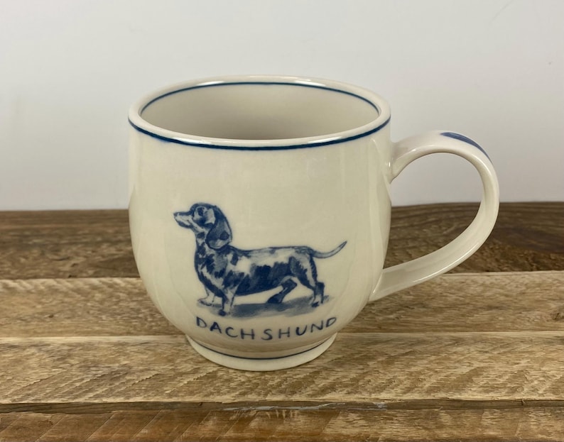 K&ouml;nnte beinhalten: Cremefarbene Keramiktasse mit einer blauen Dackel-Illustration und dem Wort "DACHSHUND" darunter. Die Tasse hat eine abgerundete Form, einen Henkel und einen blauen Rand.