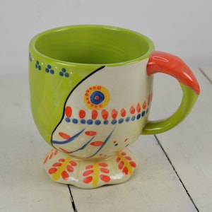 May include: A green and white ceramic mug with a colorful floral design and a red handle. The mug has a wide base and a rounded shape.