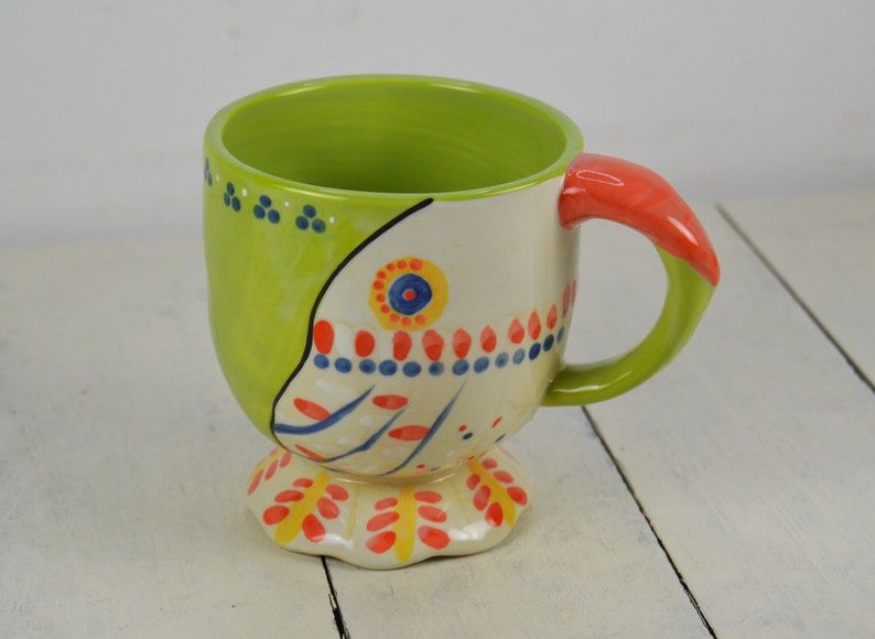 May include: A green and white ceramic mug with a colorful floral design and a red handle. The mug has a wide base and a rounded shape.