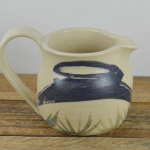 May include: A white ceramic creamer with a blue and green design. The design features a stylized bird with a long beak and wings. The creamer has a handle and a spout.