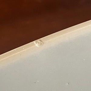 Op de afbeelding: Een close-up van een lichtgekleurd oppervlak met een bruine rand. Een klein, getextureerd, gebroken wit object bevindt zich in de rechterbovenhoek. De afbeelding is goed belicht, met focus op de details van de oppervlakken.