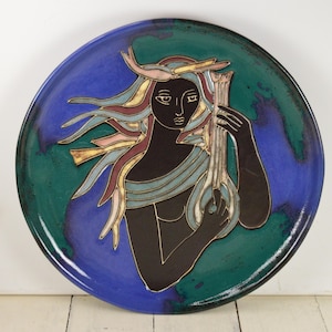 May include: A ceramic wall plate with a blue and green background featuring a stylized image of a woman with flowing hair and a musical instrument. The woman is depicted in black and gold, with colorful accents in her hair and clothing.