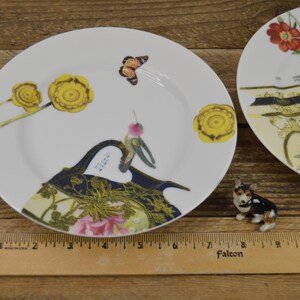 Puede incluir: Dos platos de cer&aacute;mica blanca con dise&ntilde;os florales y de mariposas. Los platos presentan un colibr&iacute;, flores amarillas y una mariposa. Los platos est&aacute;n sobre una superficie de madera.