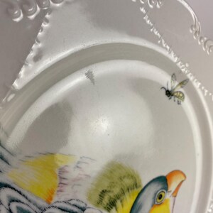 May include: White ceramic plate with a scalloped edge and a painted bird and bee design. The bird features yellow, green, and blue feathers, with an orange beak. A small bee is depicted near the edge of the plate.