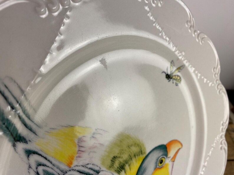 May include: White ceramic plate with a scalloped edge and a painted bird and bee design. The bird features yellow, green, and blue feathers, with an orange beak. A small bee is depicted near the edge of the plate.