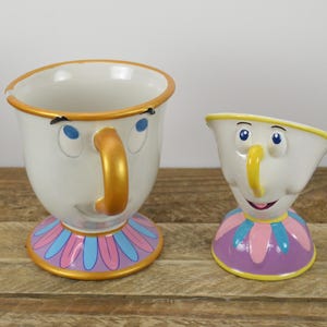 May include: Two ceramic teacups, one large and one small, with cartoon faces. The cups are white with gold trim, blue eyes, and yellow noses. The bases are decorated with pink, blue, and purple patterns.