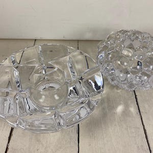 May include: Two clear glass candle holders. One is a round, segmented dish with a central candle cup. The other is a spherical, textured candle holder. Both are on a light-colored wooden surface.