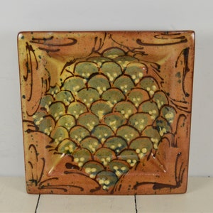 May include: A square ceramic dish with a brown, green, and yellow glaze. The dish has a raised design of overlapping scales in the center.