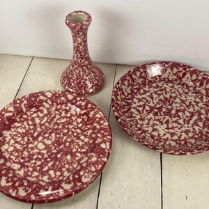 May include: A set of three ceramic pieces with a red and cream speckled pattern. The set includes a small vase and two plates, one round and one oval. The pieces are arranged on a white wooden surface.