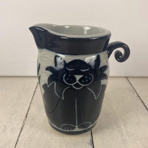 May include: A ceramic creamer with a dark blue exterior and a light gray interior. The creamer features a whimsical cat design in dark blue, with a curled handle. The creamer is approximately 5 inches tall.