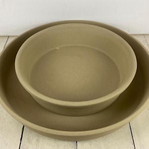 May include: Two beige stoneware baking dishes, one nested inside the other. The larger dish has a wide, shallow design, while the smaller dish is deeper. Both have rounded edges and a matte finish, suitable for baking and serving.