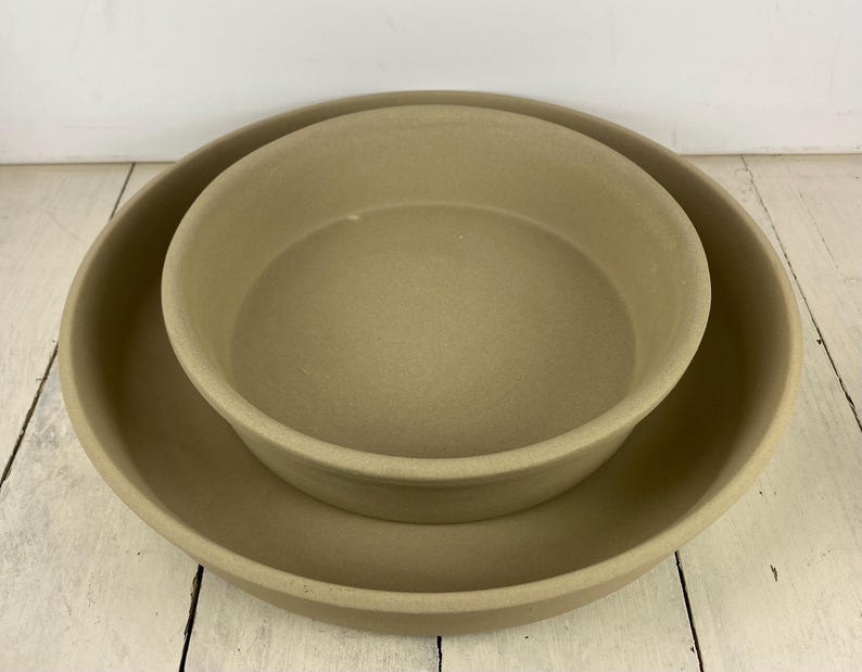 May include: Two beige stoneware baking dishes, one nested inside the other. The larger dish has a wide, shallow design, while the smaller dish is deeper. Both have rounded edges and a matte finish, suitable for baking and serving.