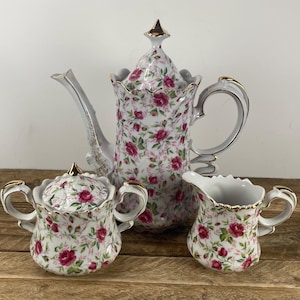 May include: A vintage porcelain tea set featuring a teapot, sugar bowl with lid, and creamer. The set is decorated with a white background and a pattern of pink roses and green leaves. Gold trim accents the edges.