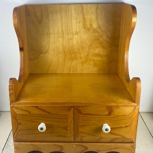 May include: A light brown wooden desktop organizer featuring a tall back panel with curved sides and a base section containing two drawers. Each drawer is equipped with a small, round, white ceramic knob centered on the front panel.