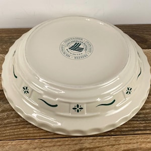 May include: Off-white ceramic pie dish with a scalloped edge and green floral and wave pattern. The bottom of the dish has a circular stamp with text that reads "Dishwasher, Microwave, Freezer, Oven Safe". Made in the U.S.A.