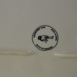 May include: A white ceramic plate with a black and white circular sticker that says "Dishwasher Safe" and "Made in China" with a small bird illustration in the center.