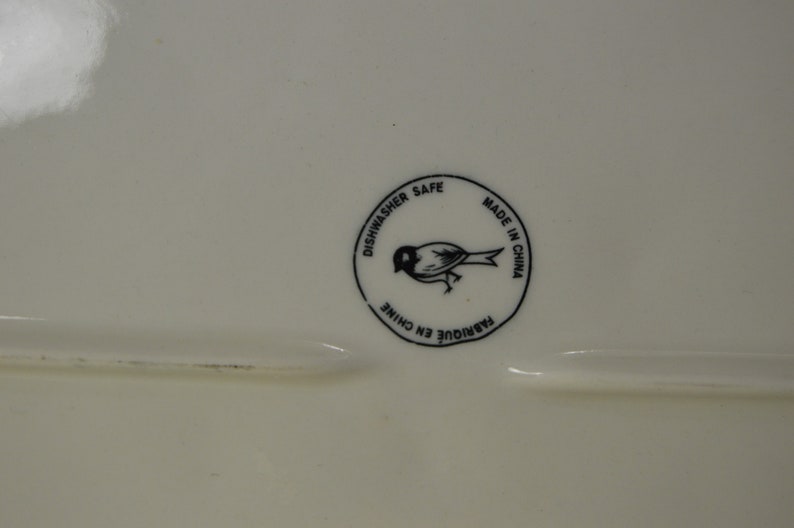 May include: A white ceramic plate with a black and white circular sticker that says "Dishwasher Safe" and "Made in China" with a small bird illustration in the center.