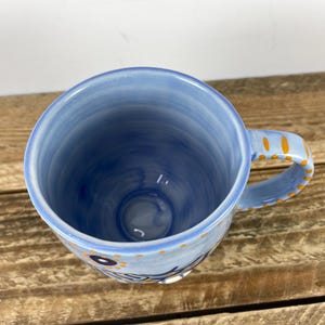 Op de afbeelding: Een blauwe keramische mok met een handvat, met een lichtblauw interieur en een donkerdere blauwe buitenkant. Het handvat is versierd met oranje stippen. De mok heeft een bloemenontwerp en het woord "Sunshine" aan de zijkant.