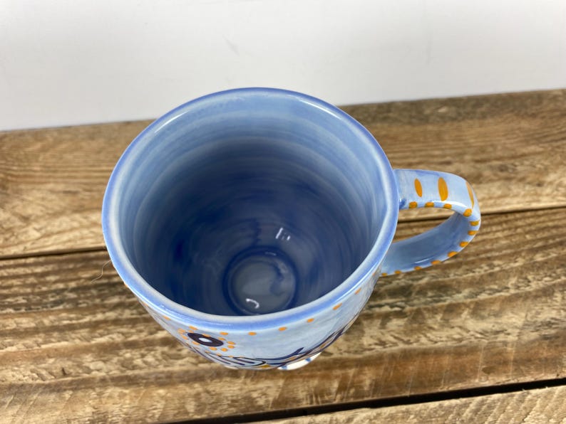 Op de afbeelding: Een blauwe keramische mok met een handvat, met een lichtblauw interieur en een donkerdere blauwe buitenkant. Het handvat is versierd met oranje stippen. De mok heeft een bloemenontwerp en het woord "Sunshine" aan de zijkant.