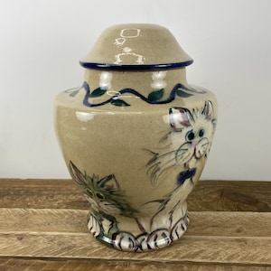 May include: Tan ceramic urn with a lid, featuring hand-painted cat illustrations. The urn has a blue rim and decorative vine accents. The cats have whimsical expressions and are painted in shades of green, purple, and blue.